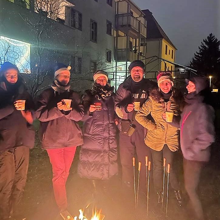 Sechs Personen stehen abends um ein Lagerfeuer vor Wohngebäuden, tragen Winterkleidung und halten Tassen in den Händen.