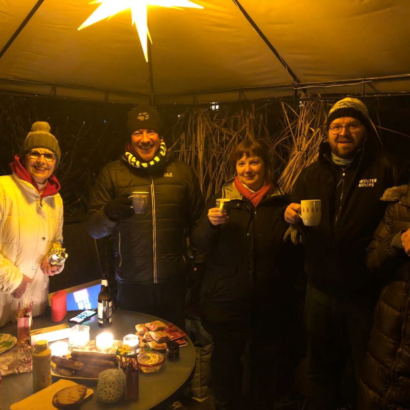 Fünf Personen in Winterkleidung stehen unter einem Zelt mit einer leuchtenden Sternlampe, halten Tassen und lächeln, vor einem Tisch mit Kerzen und Snacks