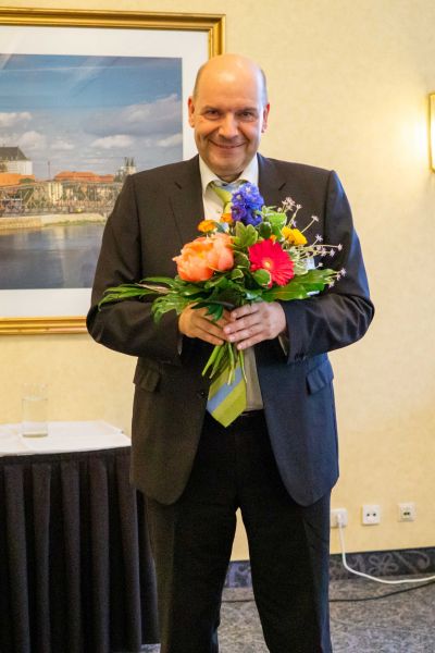 Mann im Anzug hält einen bunten Blumenstrauß und lächelt, im Hintergrund ein gerahmtes Bild einer Stadtansicht