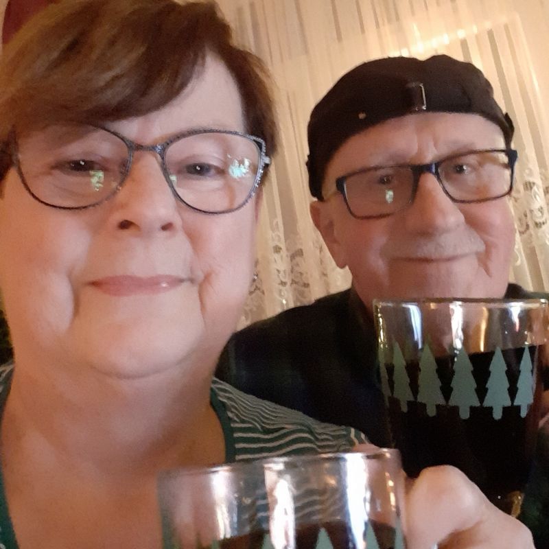 Älteres Paar mit Brillen hält Gläser mit dunklem Getränk, Hintergrund mit Gardinen