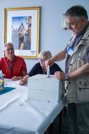 Mann mit Weste steckt einen Stimmenzettel in eine Wahlurne. Im Hintergrund sitzen zwei weitere Menschen.