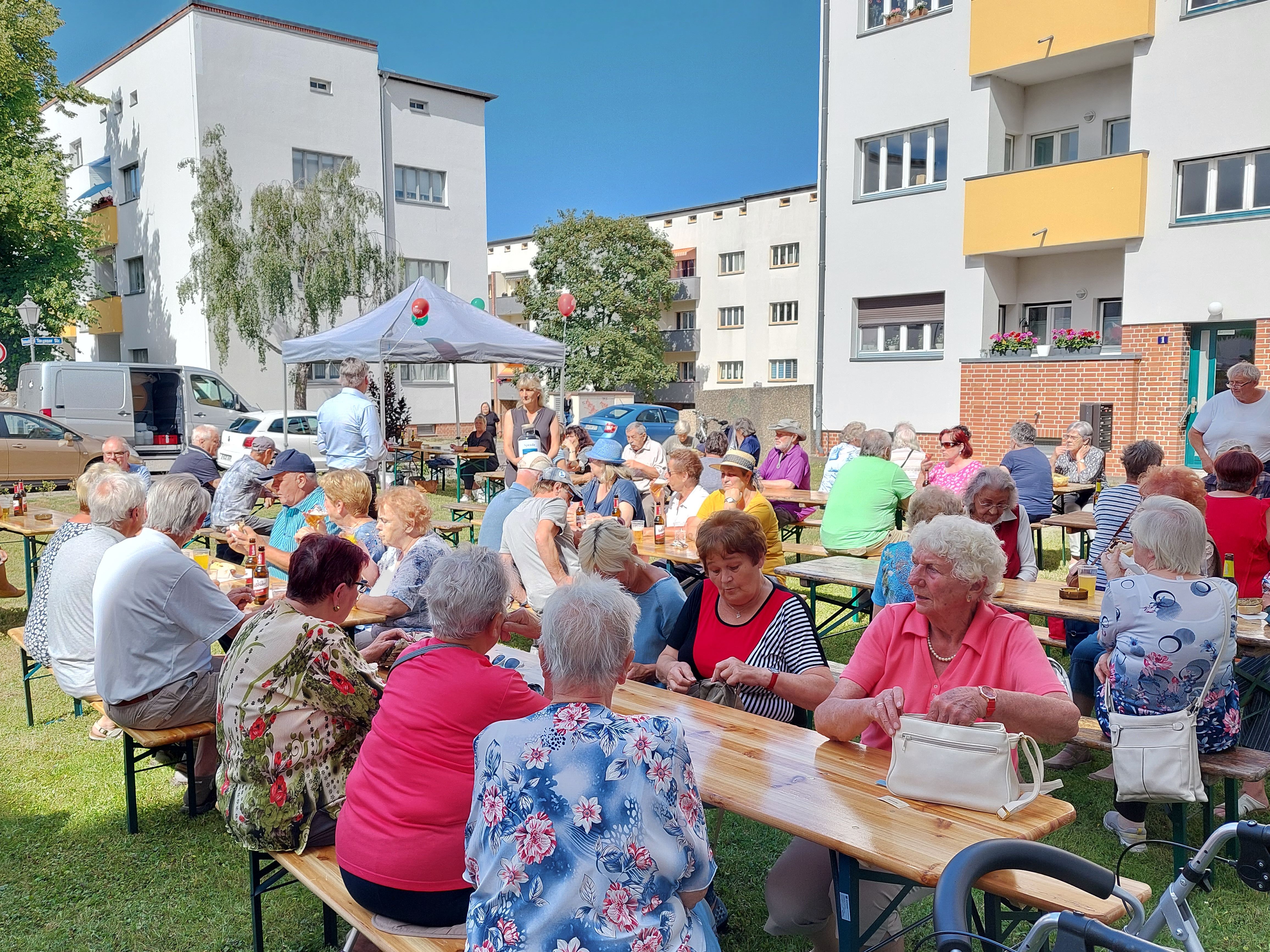 Senioren sitzen an langen Holztischen im Freien vor Wohngebäuden bei Sonnenschein