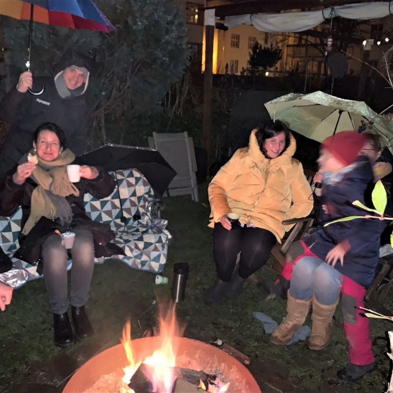 Gruppe von Menschen sitzt abends um ein Lagerfeuer im Garten, einige halten Tassen, zwei Personen halten Regenschirme, alle tragen warme Kleidung