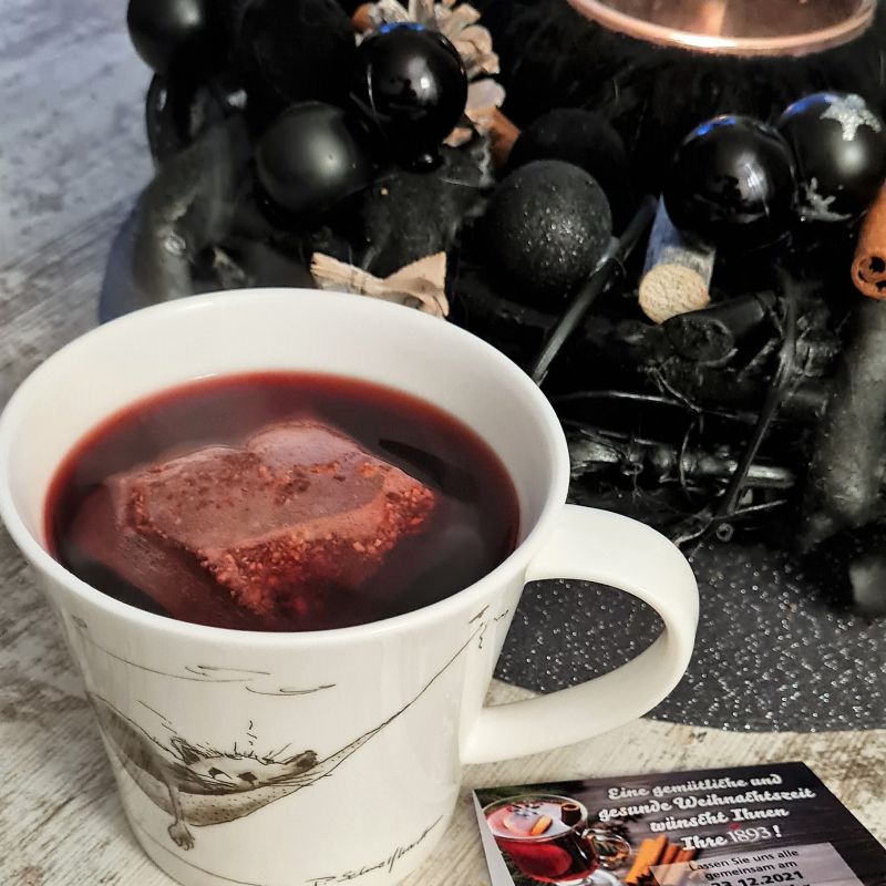 Weiße Tasse mit dampfendem roten Tee auf Holztisch, daneben Weihnachtsgrußkarte und schwarzer Adventskranz mit Kugeln und Zimtstangen