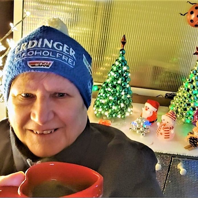 Ältere Frau mit blauer Mütze mit Aufschrift 'ERDINGER Alkoholfrei' hält rote Tasse, im Hintergrund Weihnachtsdekoration mit grünen Kugel-Weihnachtsbäumen und Figuren