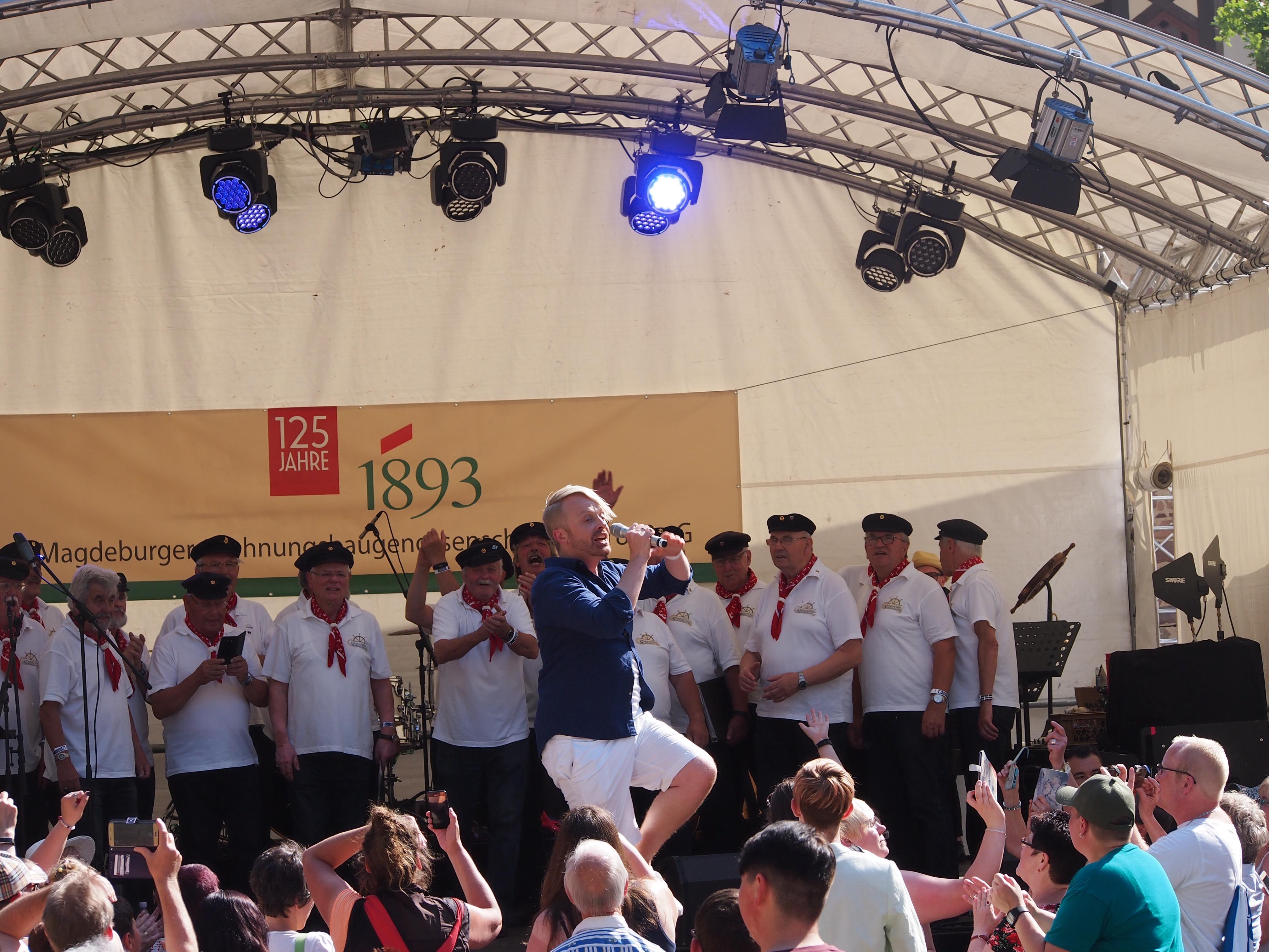 Mann steht auf der Bühne und singt, im Hintergrund ist ein Männerchor und im Vordergrund das Publikum