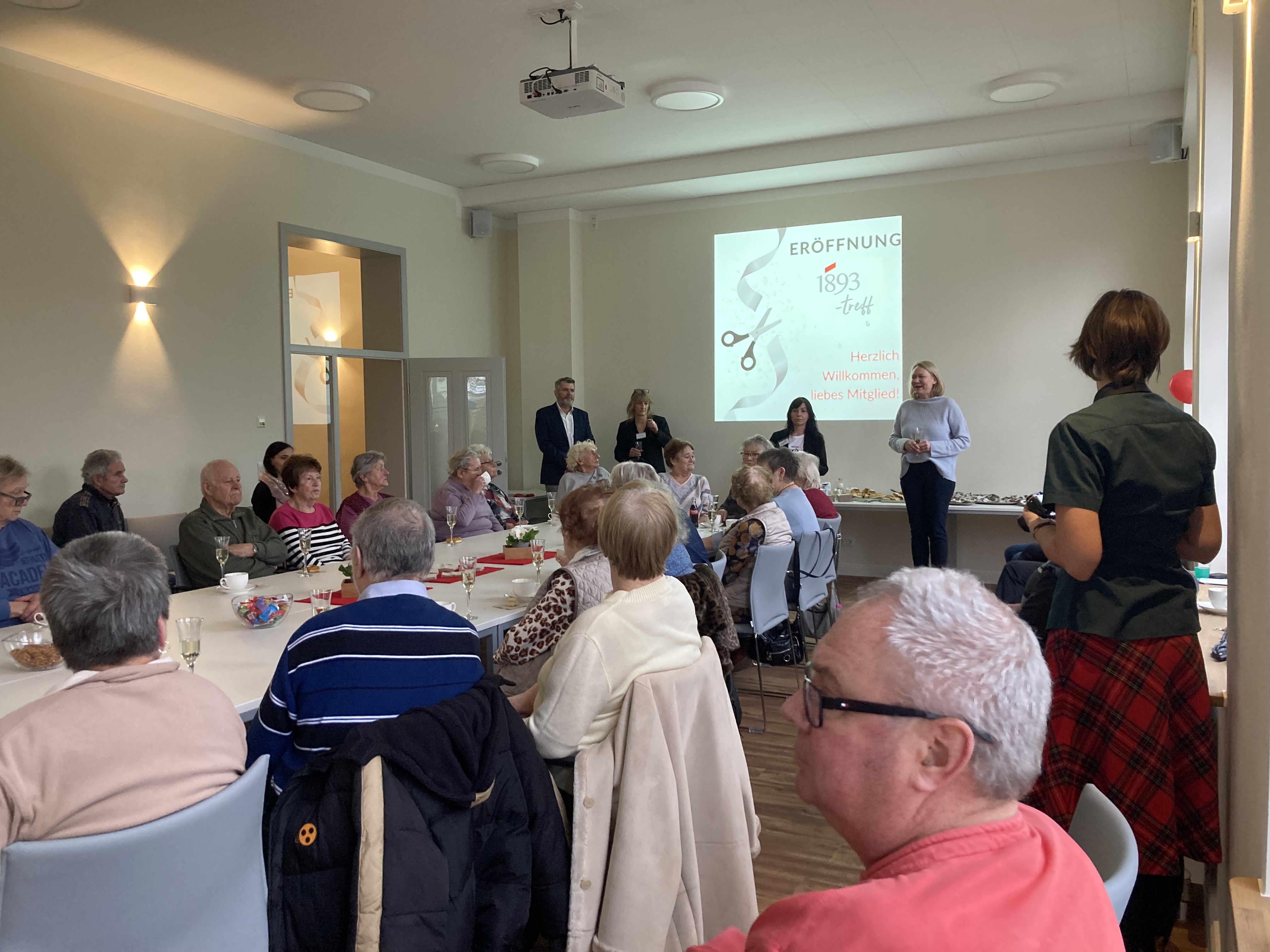Versammlung in einem hellen Raum mit Tischen, an denen ältere Menschen sitzen, während vier Personen vorne eine Präsentation halten mit dem Text 'Eröffnung 1893 trifft Herzlich Willkommen liebe Mitglieder'