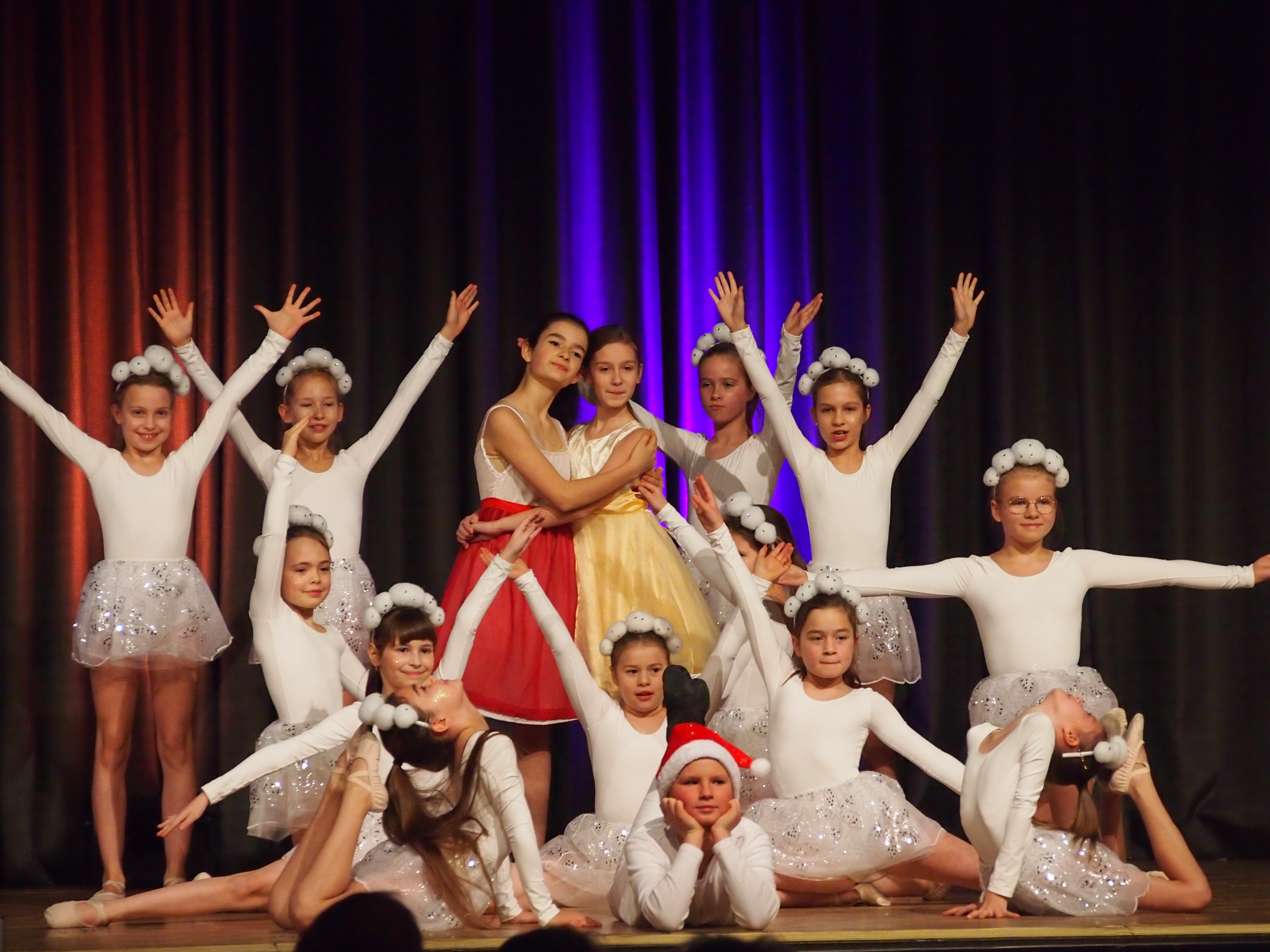 Gruppe von Mädchen in weißen Ballettkostümen mit weißen Haarschmuckbällen, zwei Mädchen in der Mitte tragen rote und gelbe Kleider, auf einer Bühne mit buntem Bühnenvorhang