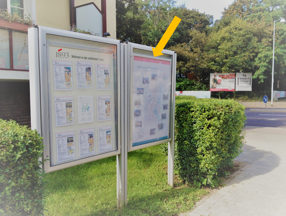 Zwei Informationsschautafeln im Freien, eine zeigt Zeitungsseiten mit der Überschrift '1893' an einem Gehweg