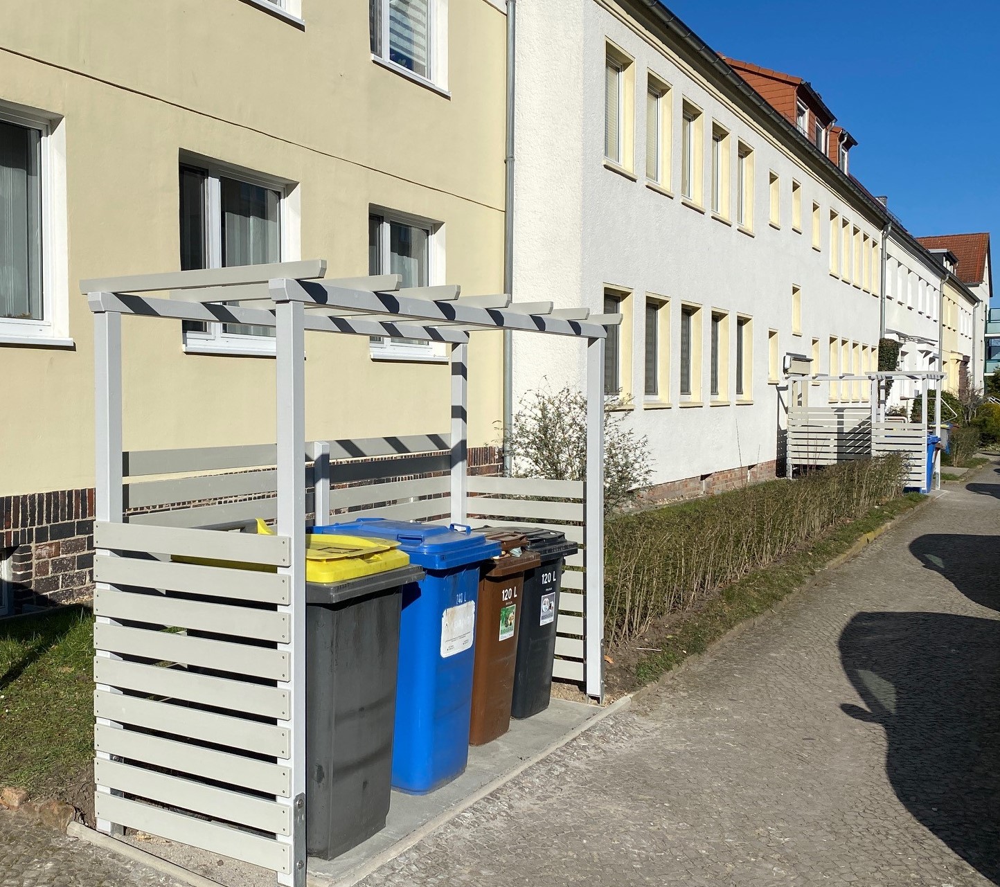 Vier Mülltonnen in verschiedenen Farben unter einem weißen Holzunterstand vor einem Wohnhaus