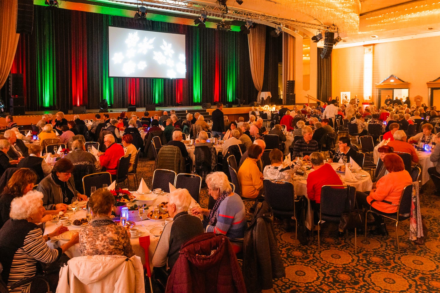 Großer festlich dekorierter Saal mit vielen Menschen an Tischen, bunte Bühnenbeleuchtung in Rot, Grün und Schwarz