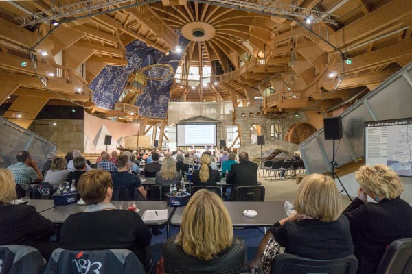 Menschen sitzen in einem Saal in Tischreihen und schauen auf eine Präsentation