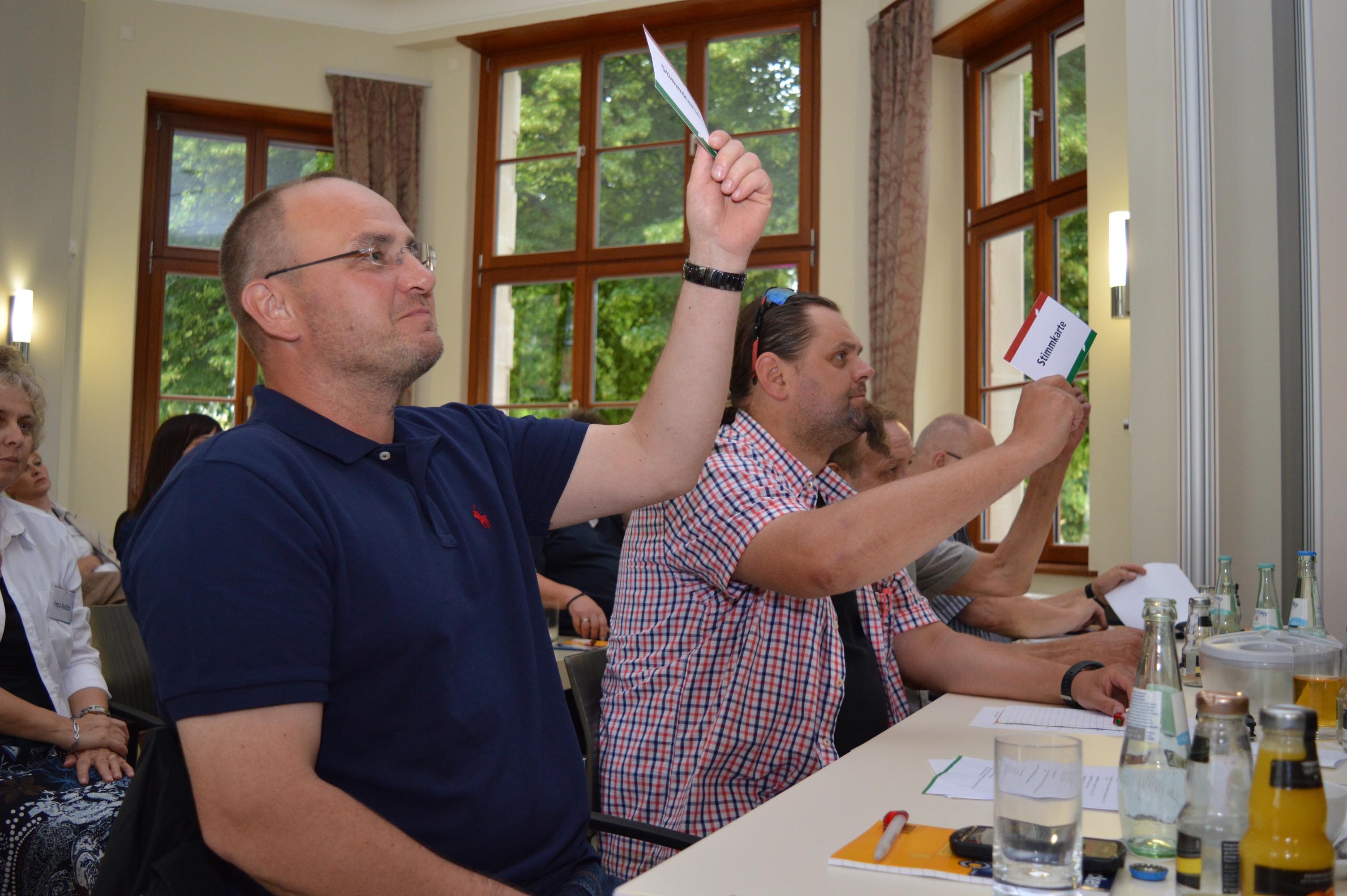 Drei Männer sitzen in einem Saal an einem Tisch und heben Stimmzettel in die Luft