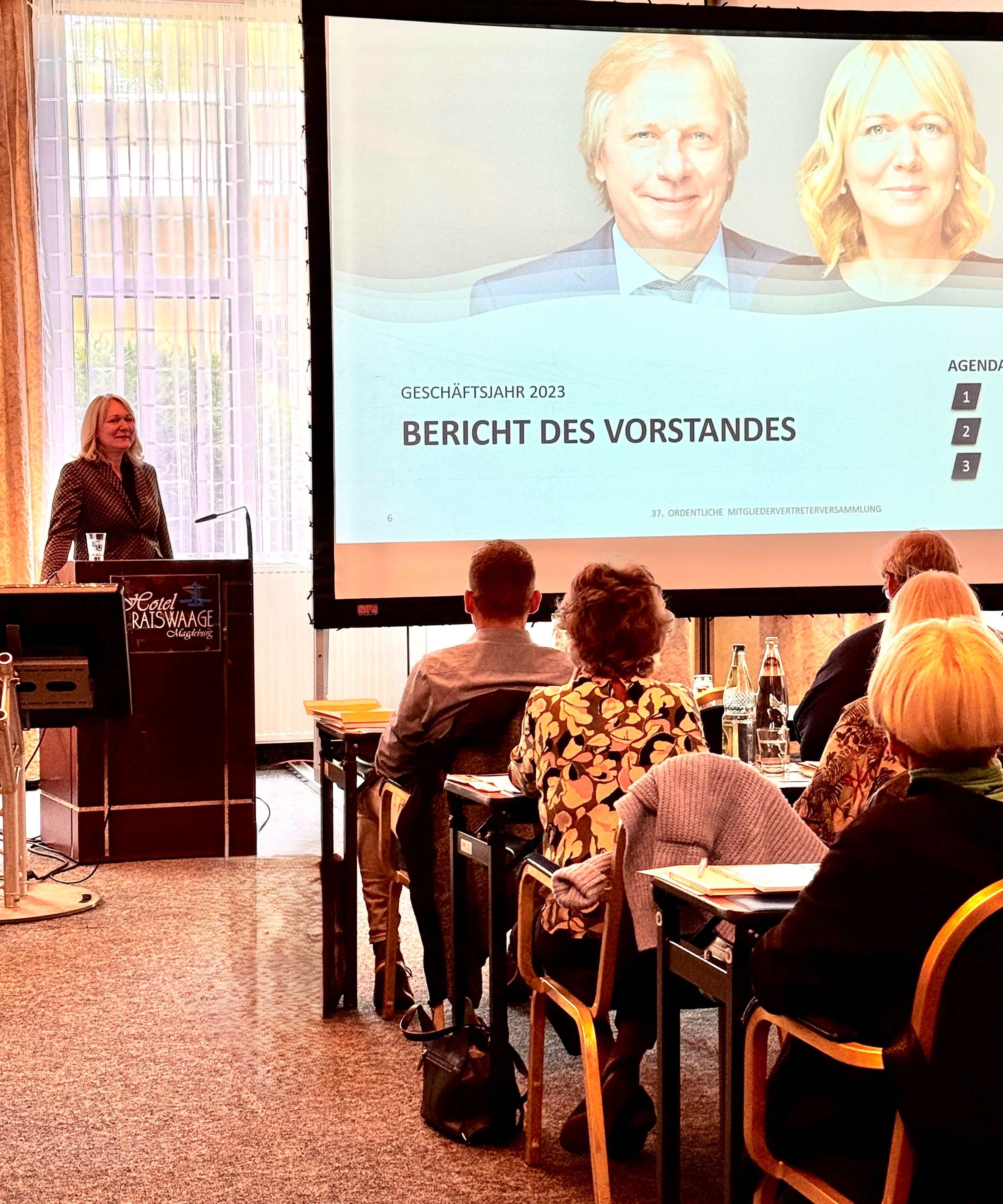 Frau hält Vortrag vor Publikum in Tagungsraum, auf Leinwand steht 'Bericht des Vorstandes, Geschäftsjahr 2023'