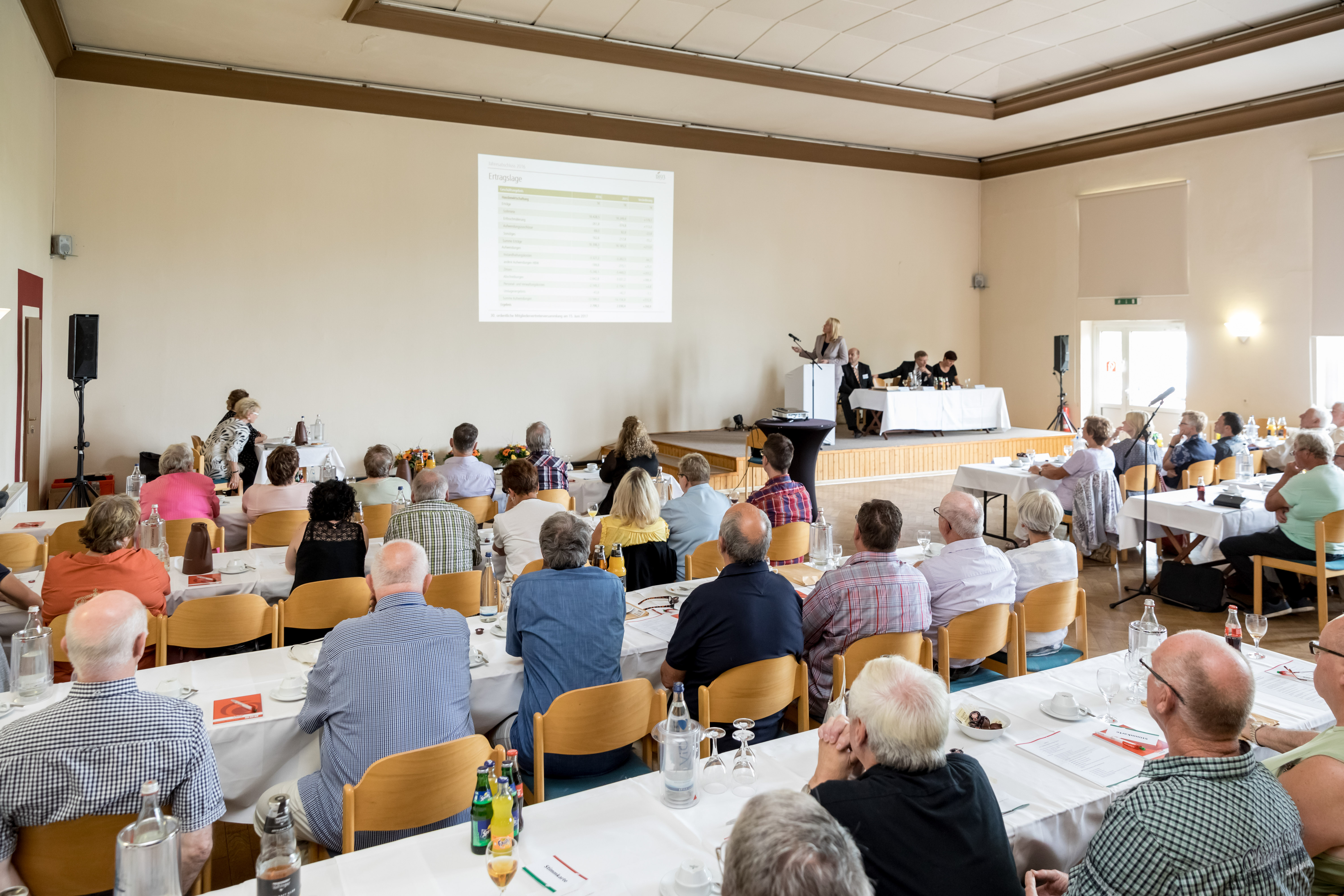 Ältere Menschen sitzen in einem Saal und hören einem Vortrag vor einer Leinwand zu