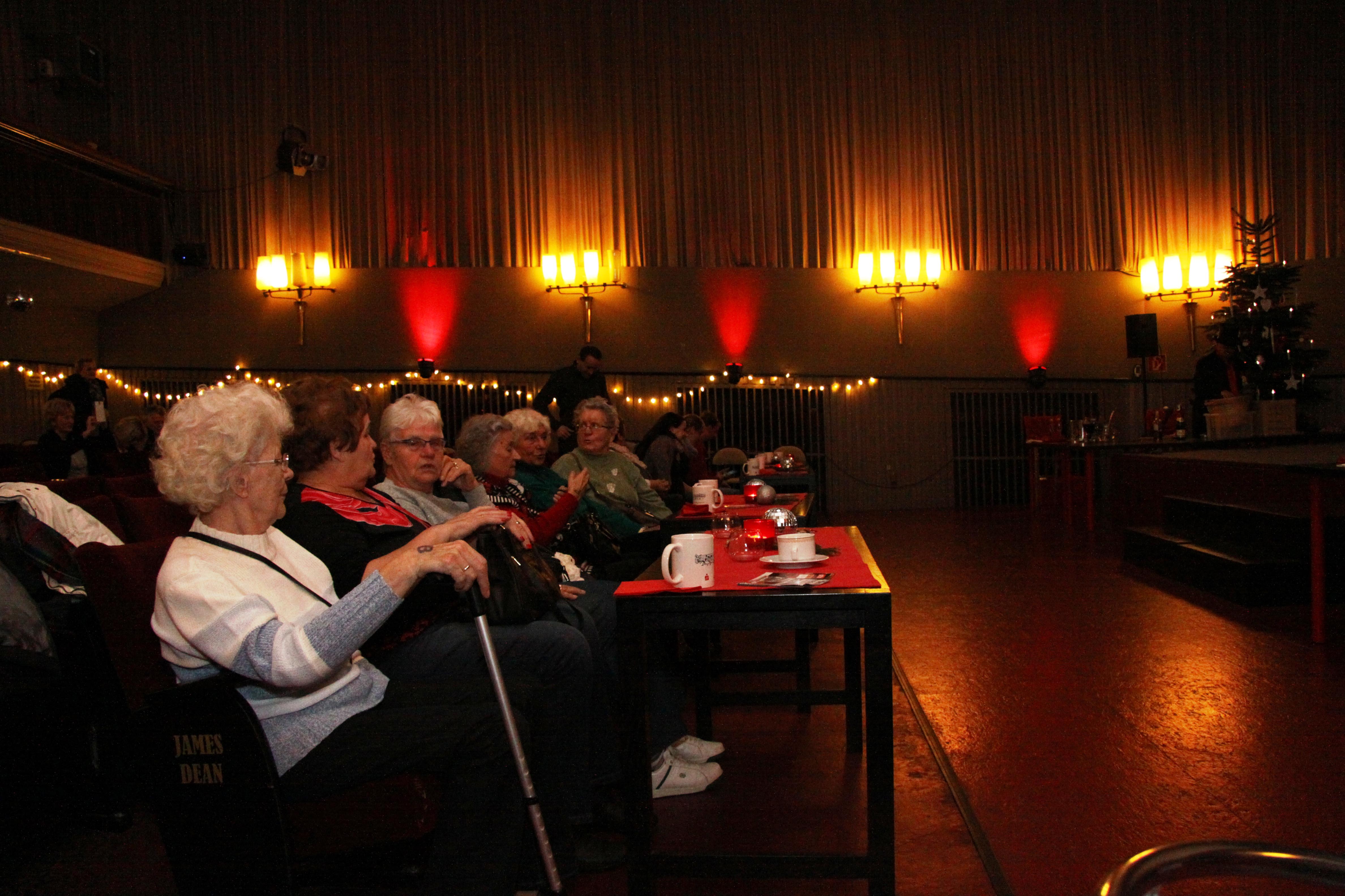 Ältere Menschen sitzen in einem Kinosaal, vor Ihnen steht ein Tisch mit Kaffee und Kuchen
