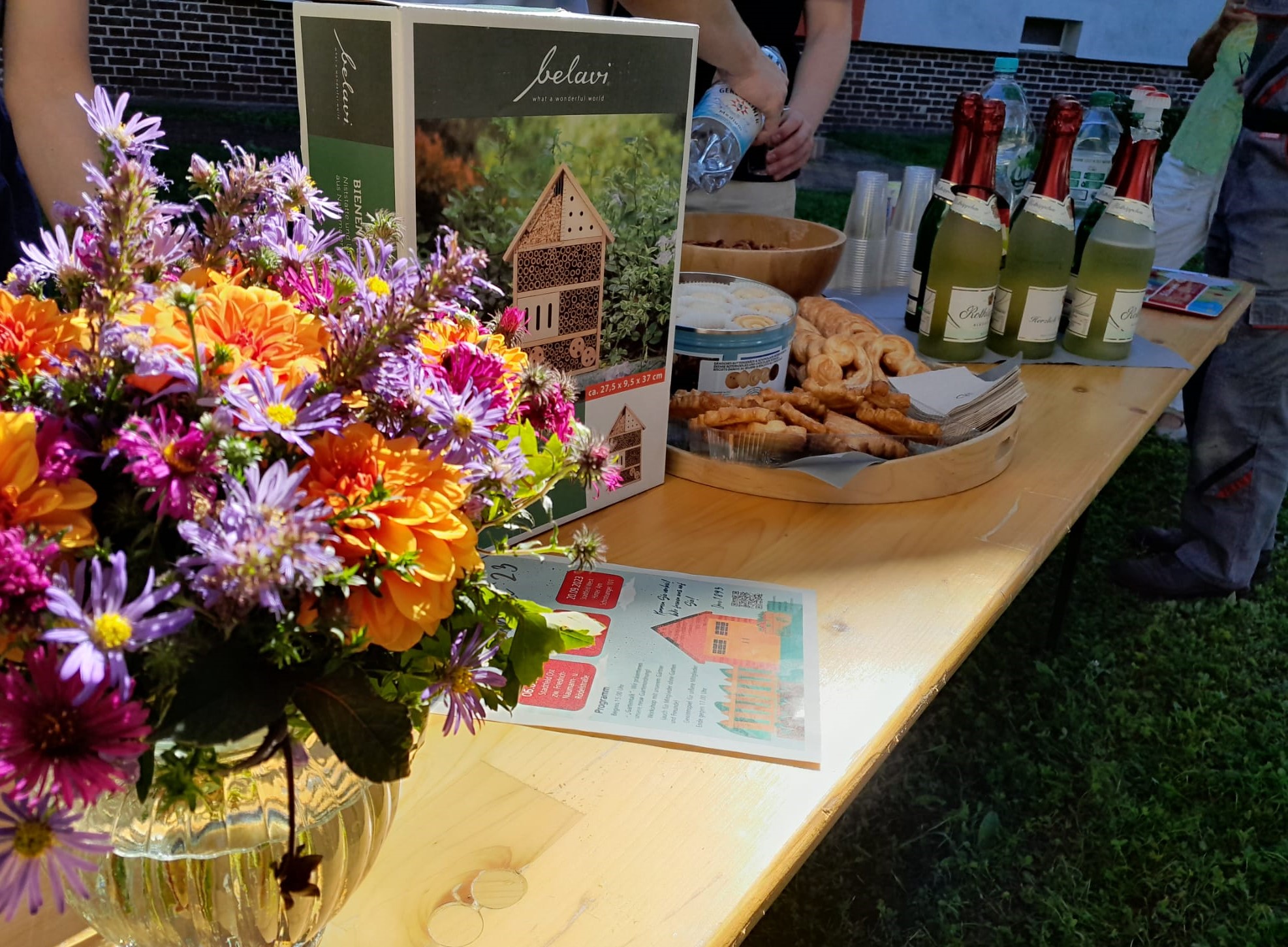 Bunter Blumenstrauß in Vase auf Holztisch mit Gebäck, Getränken und einem Insektenhotel