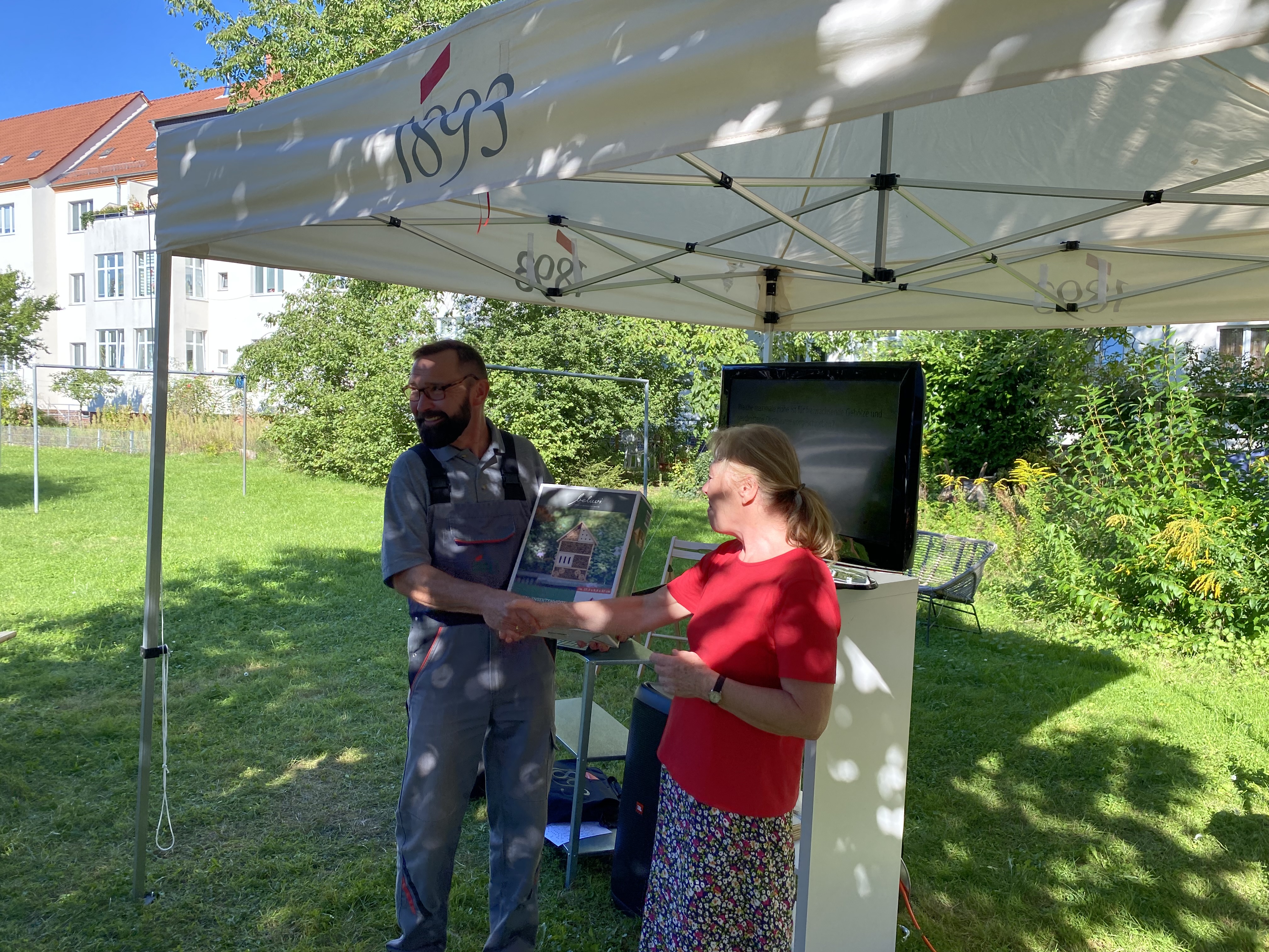 Mann in grauer Arbeitskleidung und Frau in rotem Oberteil schütteln sich unter einem weißen Pavillon die Hand, Mann hält Bienenhotel