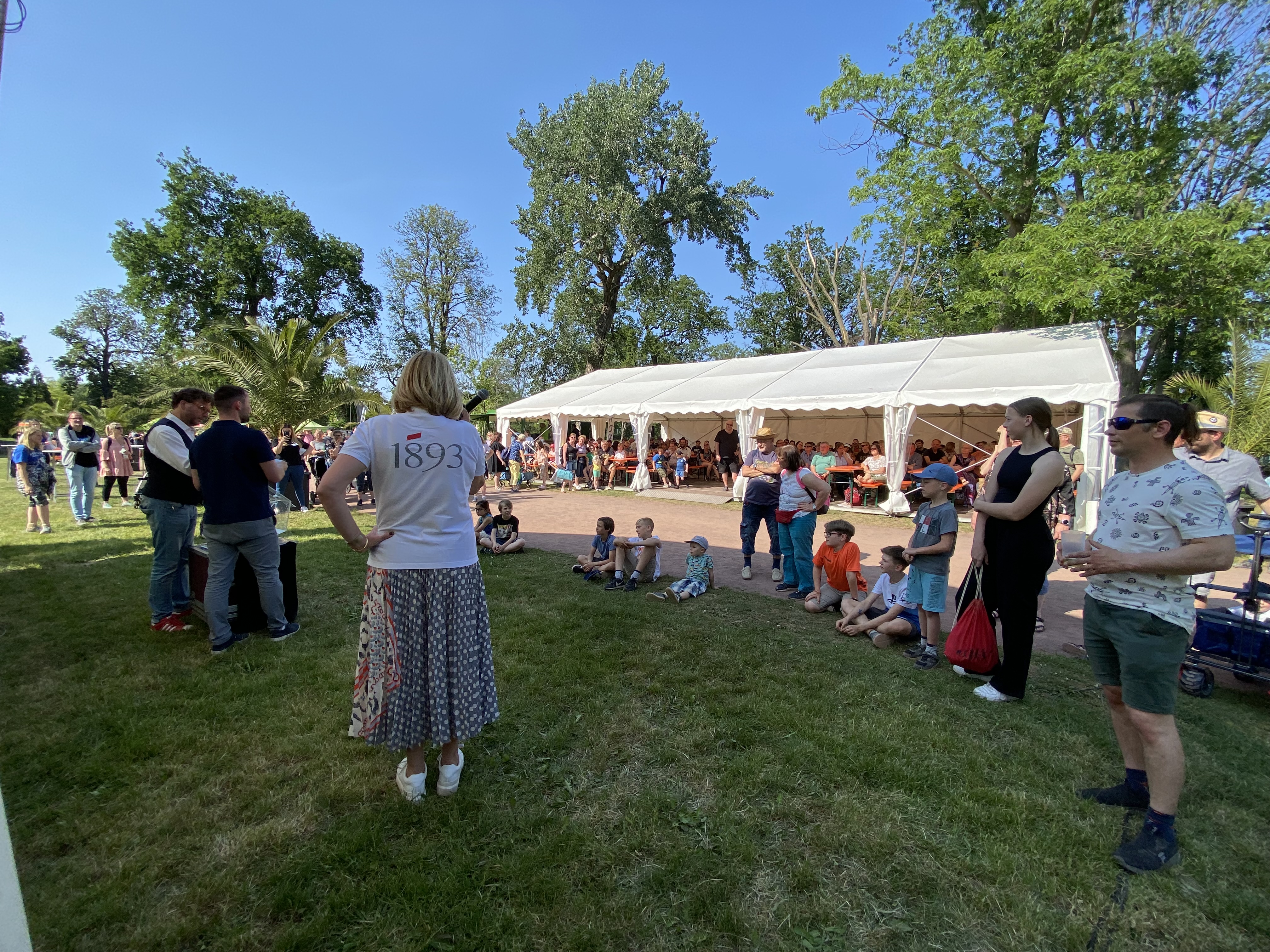 Menschen stehen auf einer Wiese vor einem weißen Festzelt, Kinder sitzen auf dem Boden