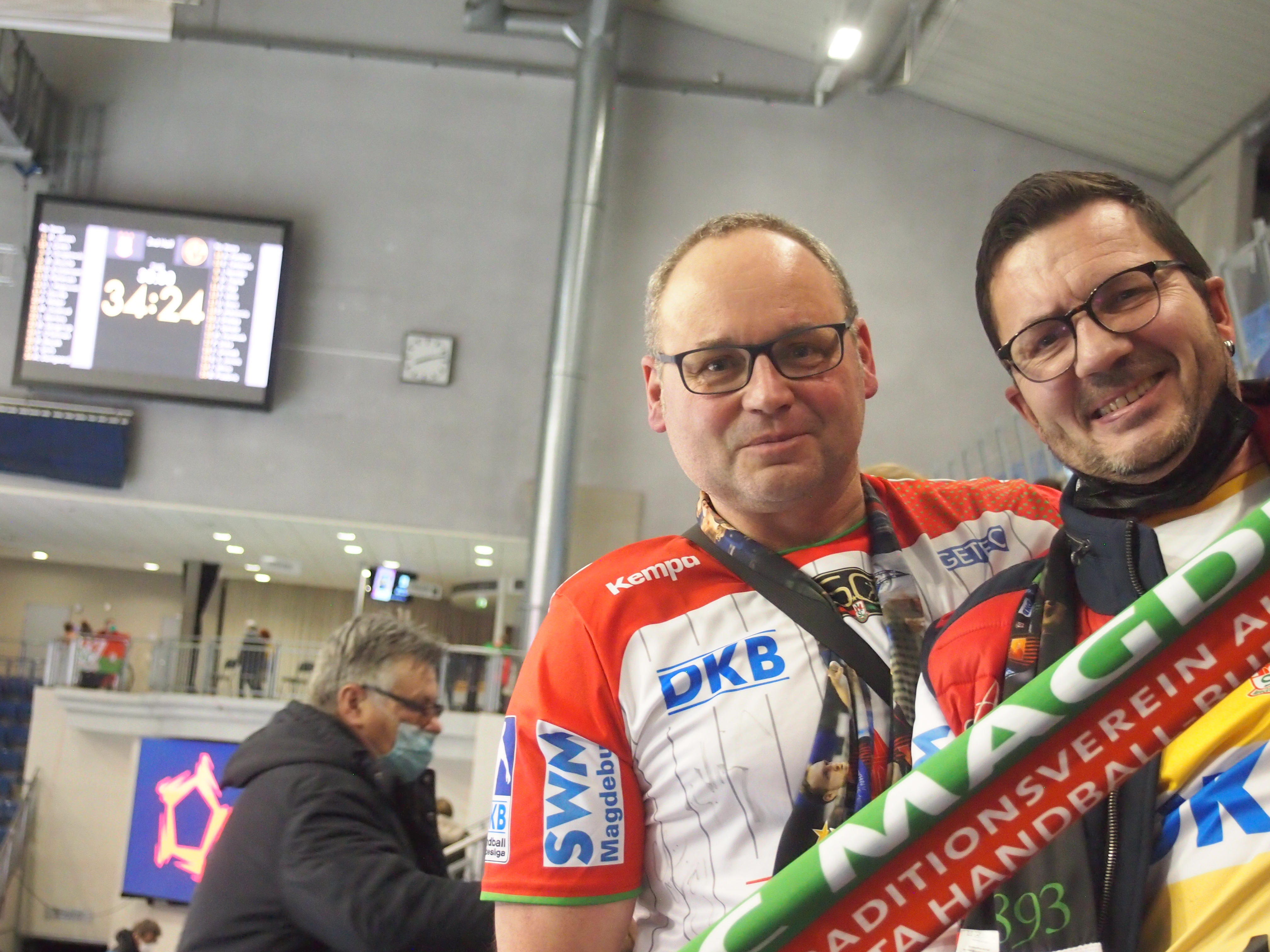 Zwei Männer lächeln in Handballtrikots mit Schal im Hintergrund ein Mann mit Maske und eine Anzeigetafel