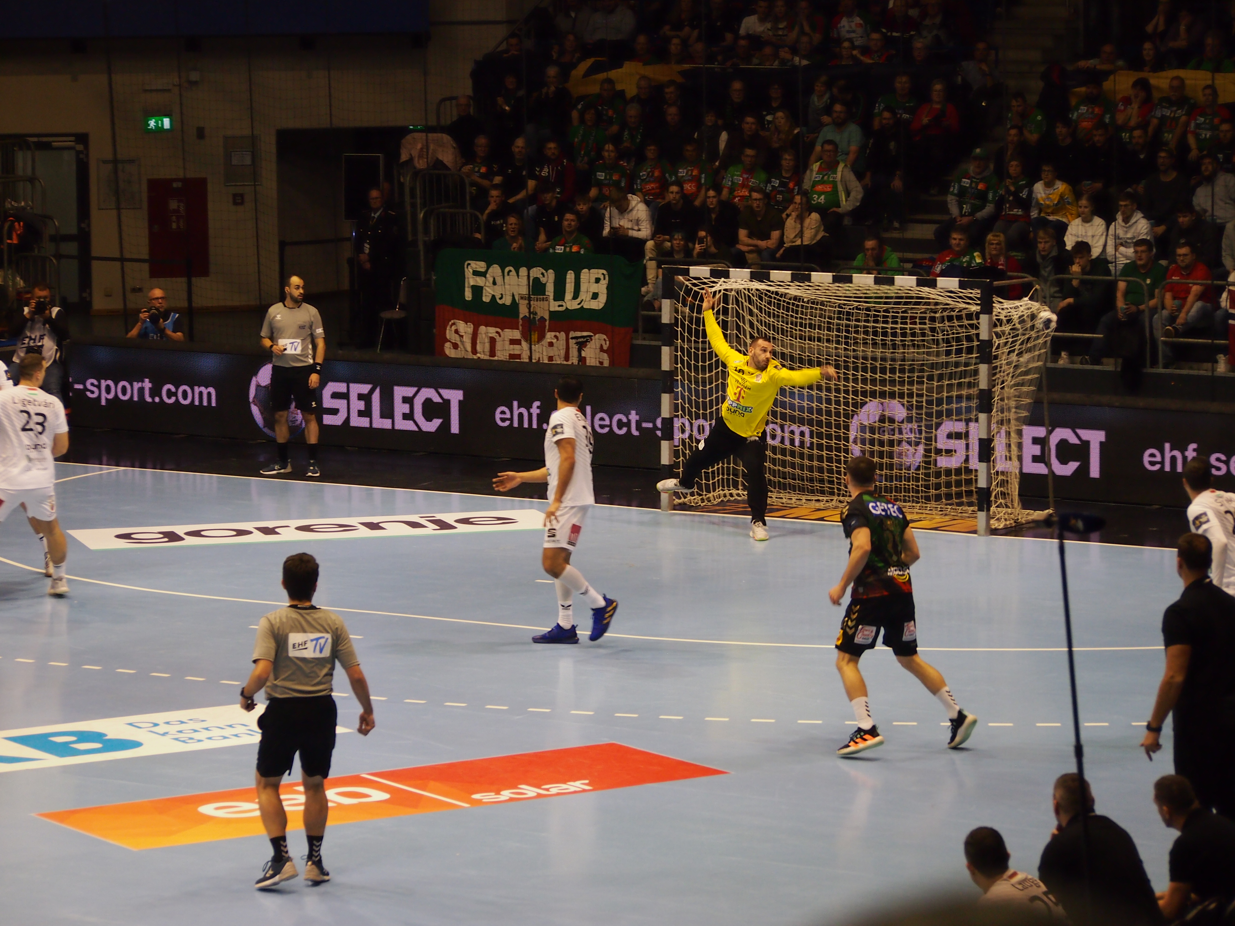 Handballtorwart in gelbem Trikot springt zum Ball vor dem Tor, Spieler in weißen und schwarzen Trikots auf dem Spielfeld, Zuschauer im Hintergrund