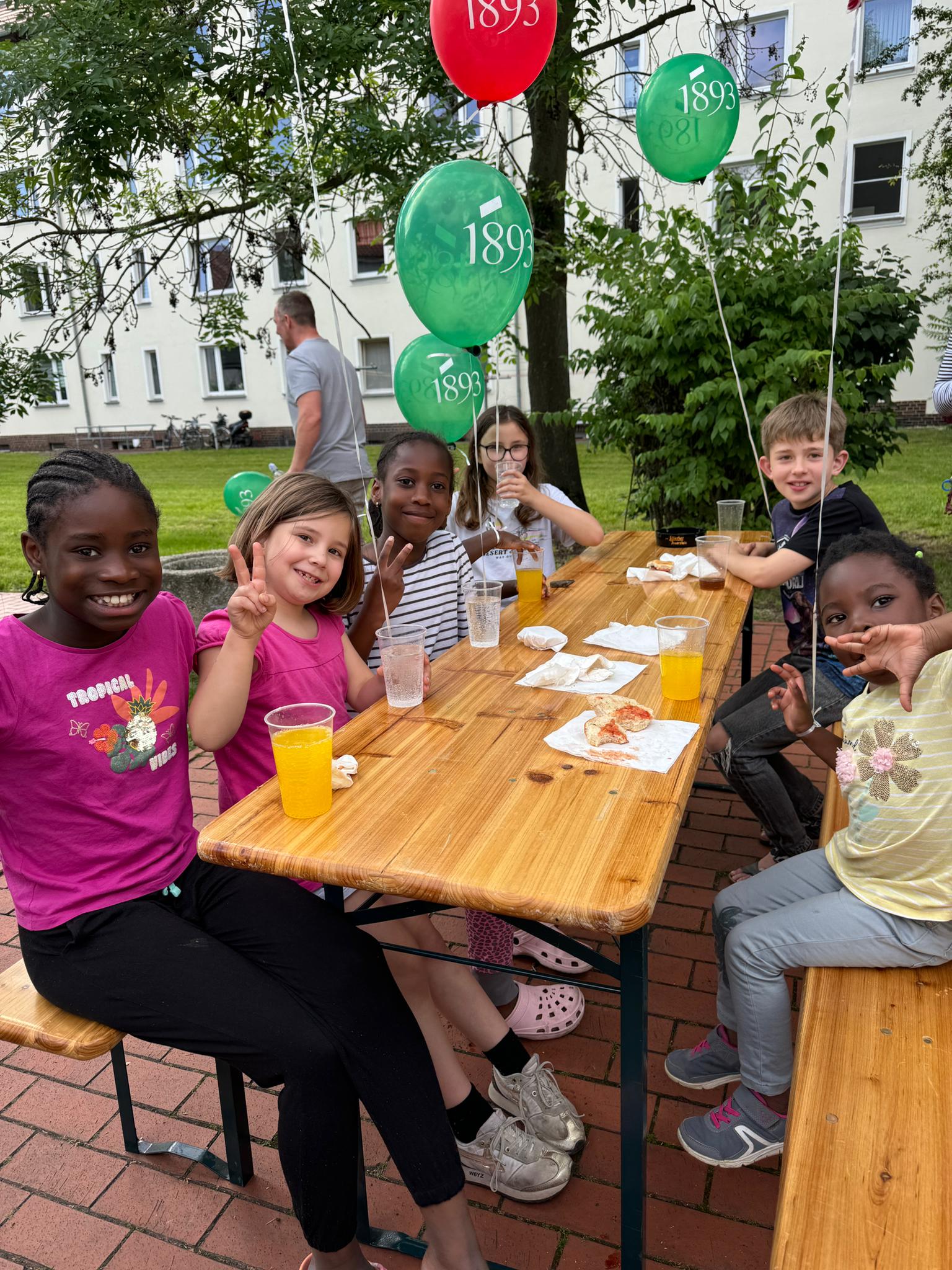 Sechs Kinder sitzen an einem Holztisch im Freien, essen und trinken, umgeben von grünen und roten Luftballons mit der Aufschrift 1893