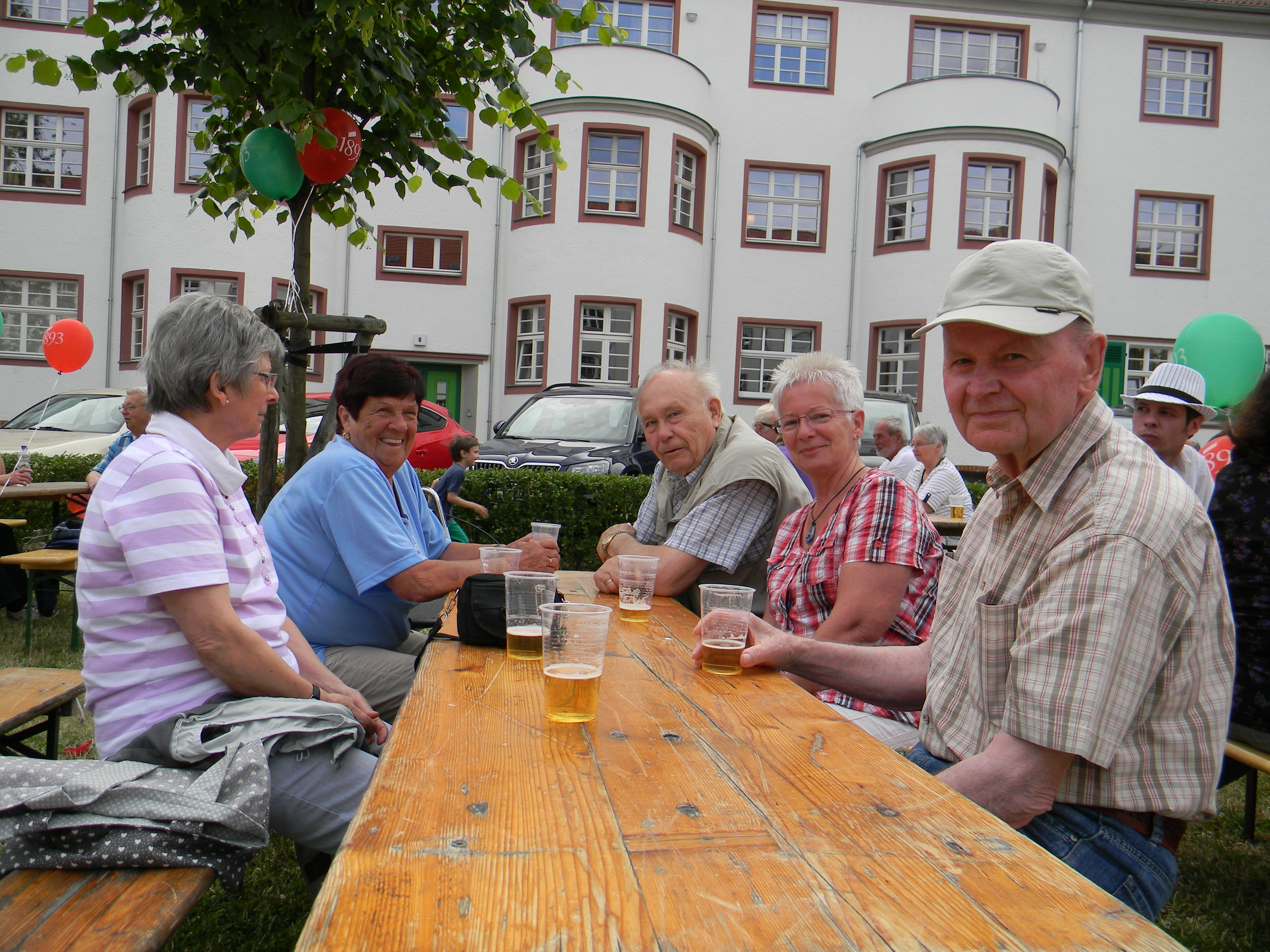 Fünf Menschen sitzen an einer Bierbank vor einem Wohnhaus und lachen in die Kamera