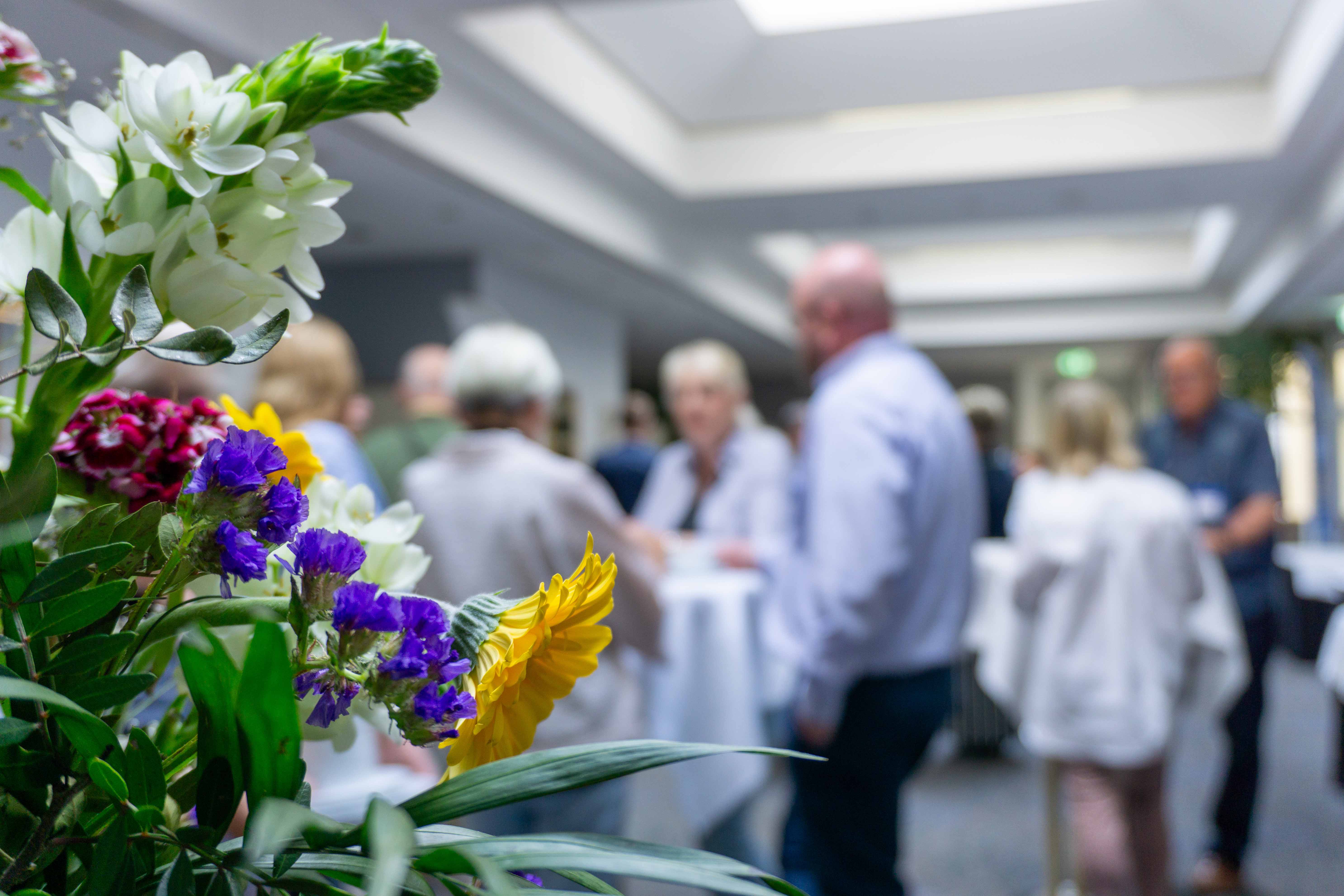 Bunte Blumen im Vordergrund, unscharfe Menschen in einem hellen Raum mit Tischen im Hintergrund