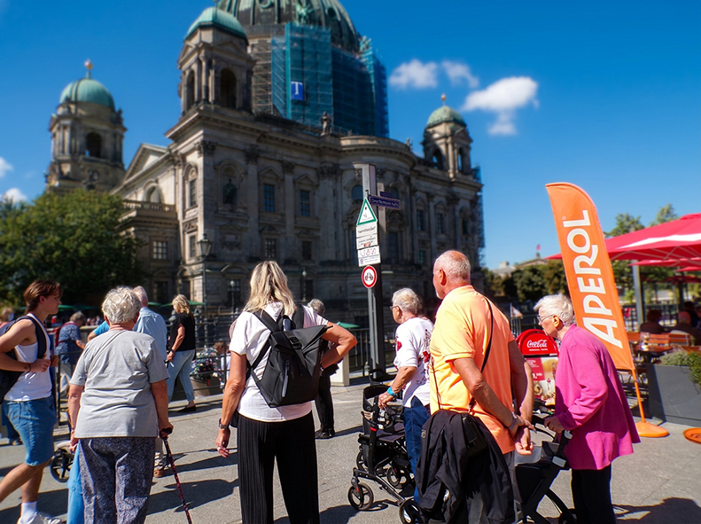 Eine Reisegruppe steht auf einem Platz vor dem Berliner Dom. Einige Teilnehmende nutzen Rollatoren oder Gehhilfen. Im Hintergrund erhebt sich die historische Kathedrale mit ihrer markanten Kuppel, die teilweise von einem Gerüst umgeben ist. Die Gruppe fol