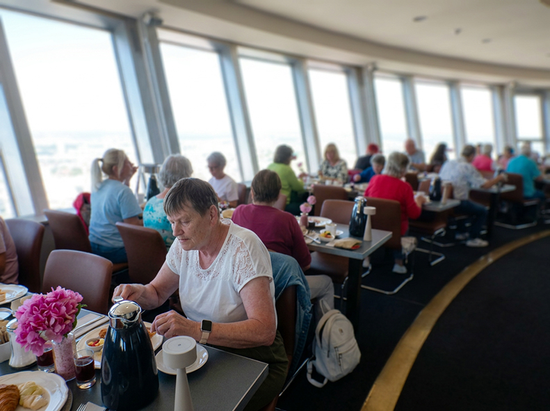 Im Restaurantbereich des Berliner Fernsehturms sitzen zahlreiche Besucher an gedeckten Tischen entlang einer großen Fensterfront. Die bodentiefen Fenster bieten einen weiten Ausblick über die Stadt. Auf den Tischen stehen Kaffee, Geschirr und Frühstückssp