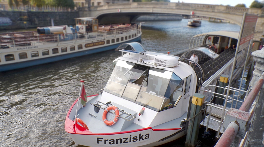 Ein weißes Ausflugsschiff mit dem Namen „Franziska“ liegt an einem Anleger auf einem Berliner Wasserweg. Im Hintergrund fahren weitere Fahrgastschiffe auf dem Fluss, während eine Steinbrücke das Gewässer überspannt. Das Schiff verfügt über große Fenster u