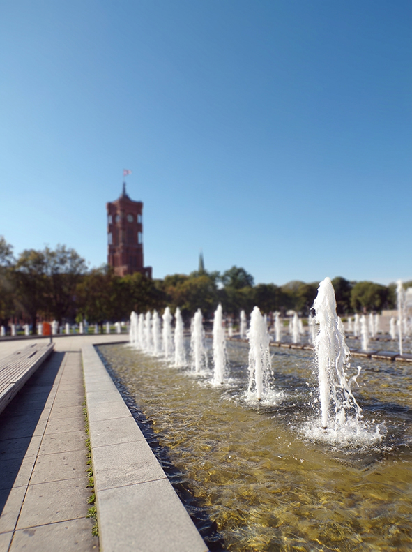 Mehrere Wasserfontänen steigen aus einem flachen rechteckigen Becken auf. Im Vordergrund ist eine einzelne Fontäne scharf zu erkennen, während die weiteren Fontänen in einer Reihe nach hinten verlaufen. Links befindet sich ein gepflasterter Weg mit Sitzmö