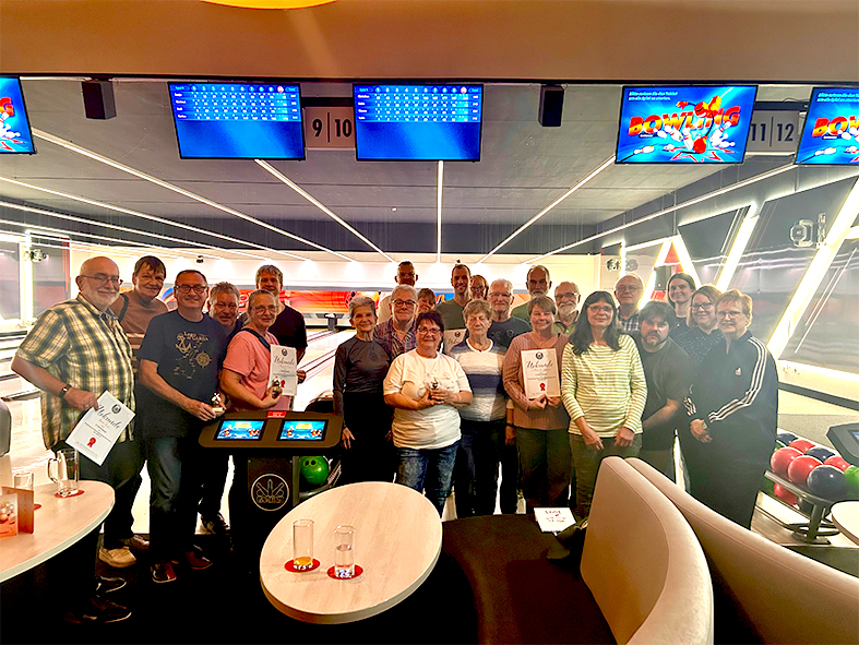 Gruppenfoto in einer Bowlinghalle. Rund zwei Dutzend Teilnehmer stehen vor den Bowlingbahnen, einige halten Urkunden und kleine Preise in den Händen. Im Hintergrund sind die Bowlingbahnen sowie digitale Anzeigetafeln zu sehen. Im Vordergrund befinden sich