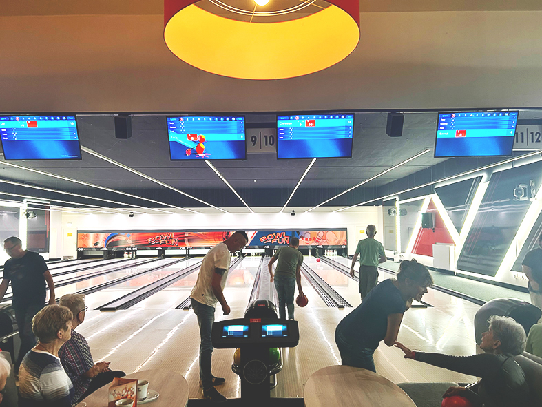 Mehrere Teilnehmer eines Bowling-Nachmittags befinden sich an den Bowlingbahnen einer modernen Bowlinghalle. Einige Personen bereiten einen Wurf vor, während andere an Tischen sitzen und das Spiel verfolgen. Über den Bahnen hängen digitale Anzeigetafeln.