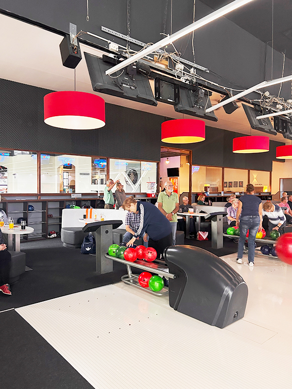 Mehrere Personen spielen Bowling in einer modernen Bowlinghalle. Im Vordergrund befinden sich rote und grüne Bowlingkugeln auf der Kugelrückgabe. Im Hintergrund sitzen weitere Teilnehmer an Tischen entlang der Bahnen. Rote Deckenleuchten prägen das Ersche