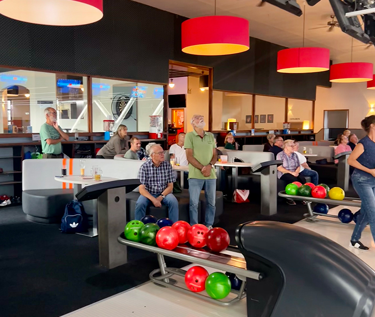 Teilnehmer eines Bowling-Nachmittags sitzen und stehen in den Sitzbereichen einer Bowlinghalle. Im Vordergrund befinden sich Bowlingkugeln auf der Kugelrückgabe. Mehrere Personen blicken in Richtung der Bowlingbahnen, während dort gespielt wird. Über den
