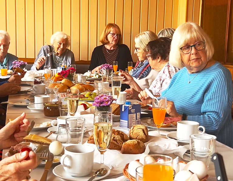 Teilnehmende eines Mieterfrühstücks sitzen an einer festlich gedeckten Tafel mit Brötchen, Eiern, Obst, Kaffee und Saftgetränken.