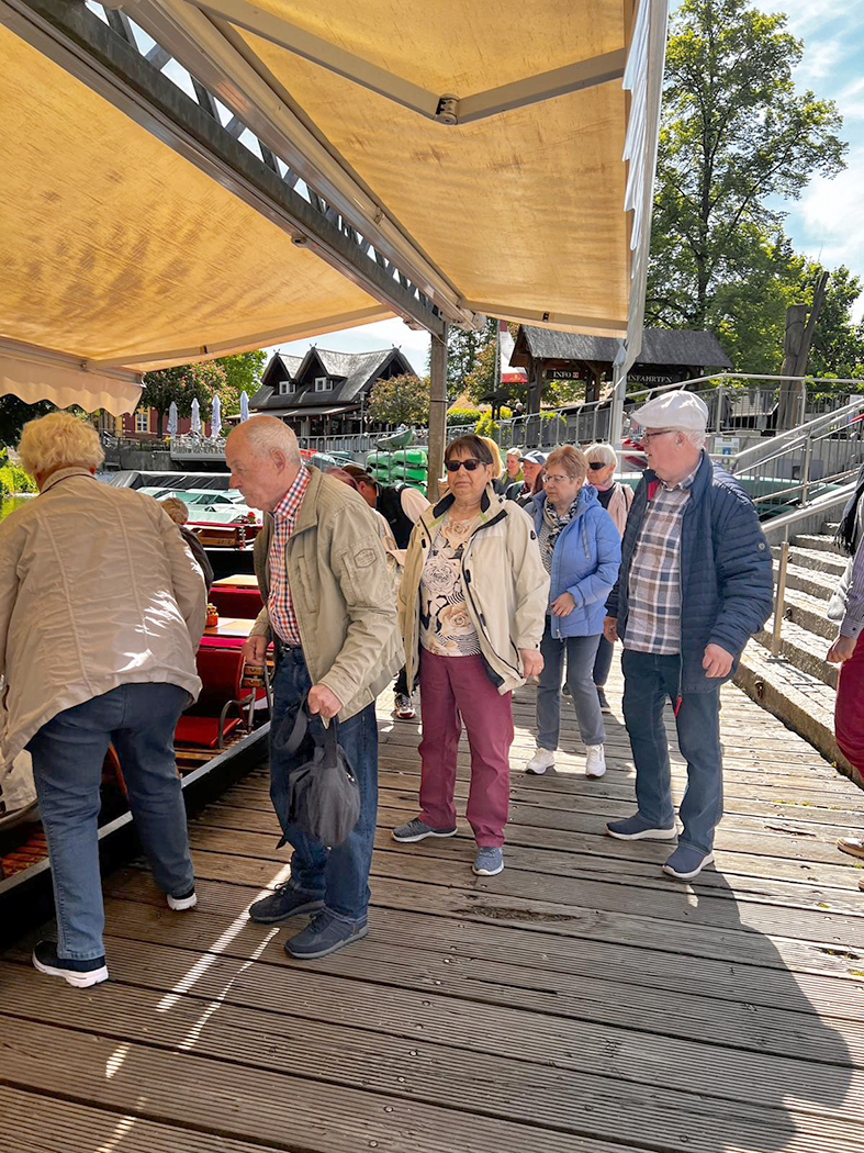 Auf einem hölzernen Steg bewegen sich mehrere Personen in Richtung eines flachen Kahns. Links im Bild steigt bereits eine Person vorsichtig in das Boot ein. Direkt dahinter warten weitere Teilnehmer auf das Boarding.