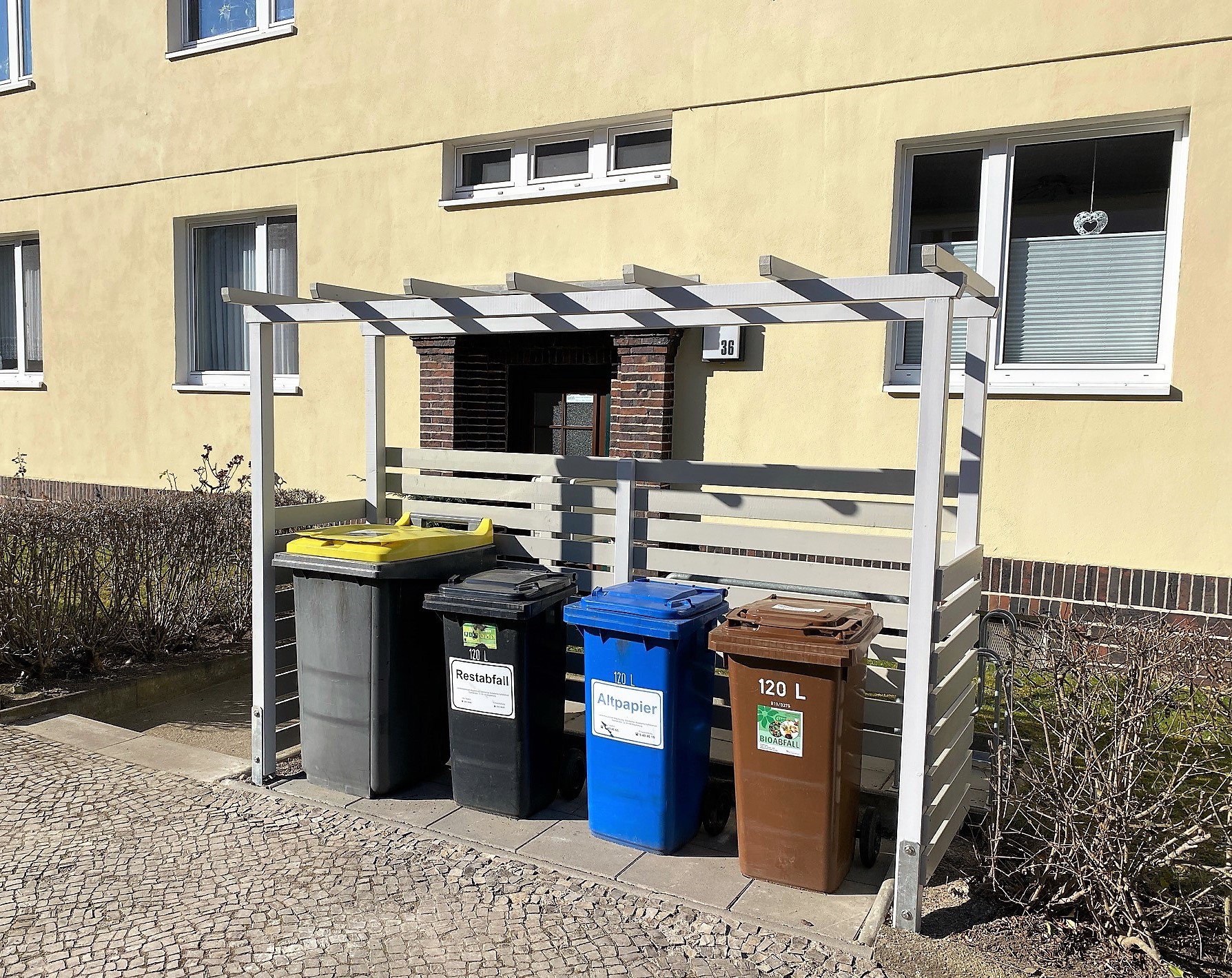 Müllplätze in der Zackelbergstraße Vier Mülltonnen unter einem weißen Holzunterstand vor einem gelben Wohnhaus mit Fenstern, beschriftet für Restabfall, Altpapier und Bioabfall