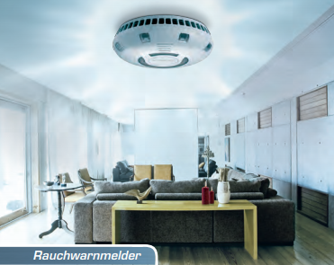 Rauchwarnmelder an der Decke eines hellen Wohnzimmers mit grauem Sofa und großem Fenster