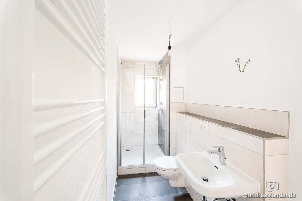 Schmales, modernes weißes Badezimmer mit Dusche, Waschbecken und Toilette auf grauem Bodenfliesen