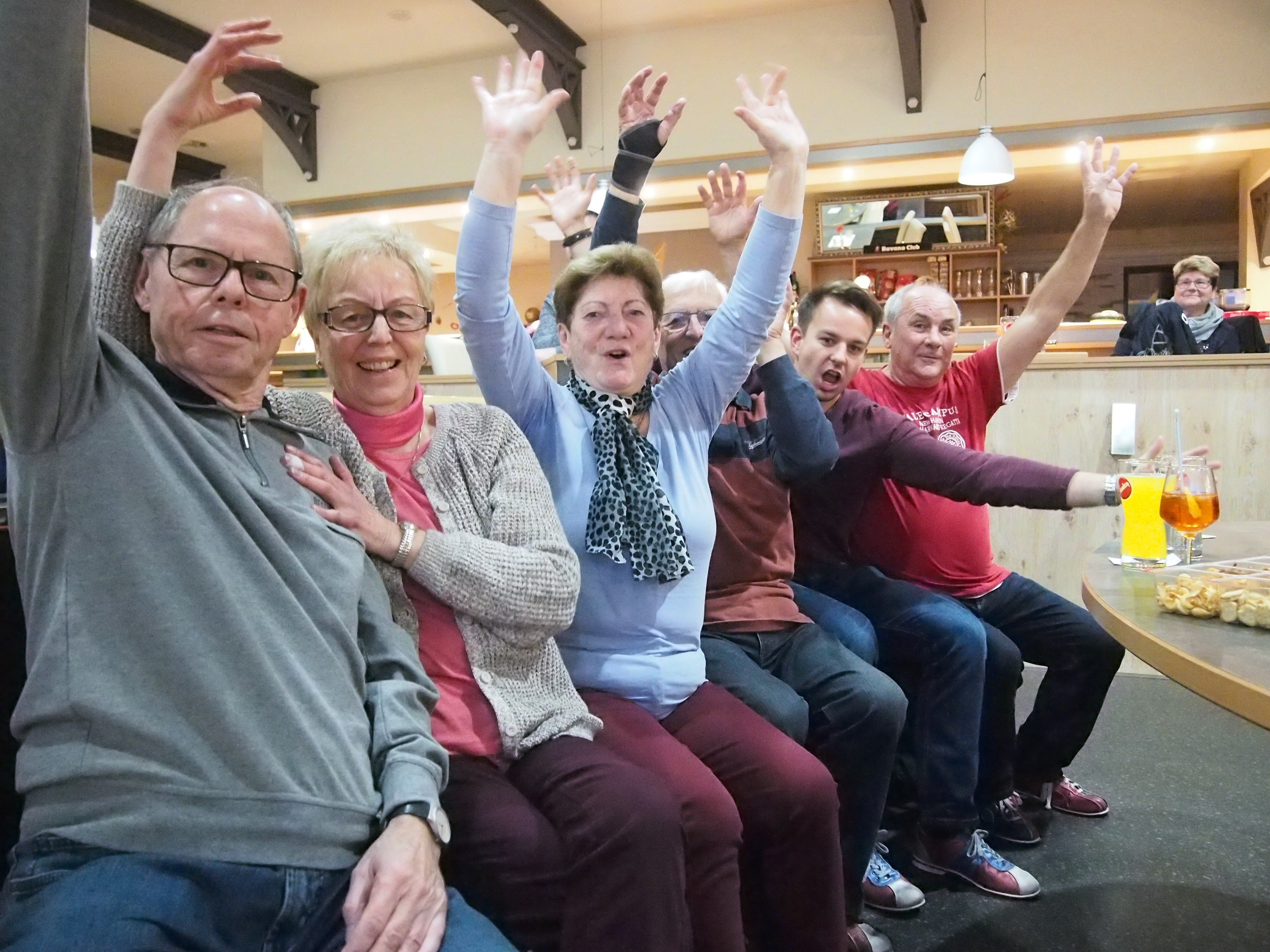 Sechs Menschen sitzen auf einer Bank in einer Bowlingbahn und jubeln mit gehobenen Armen in die Kamera.