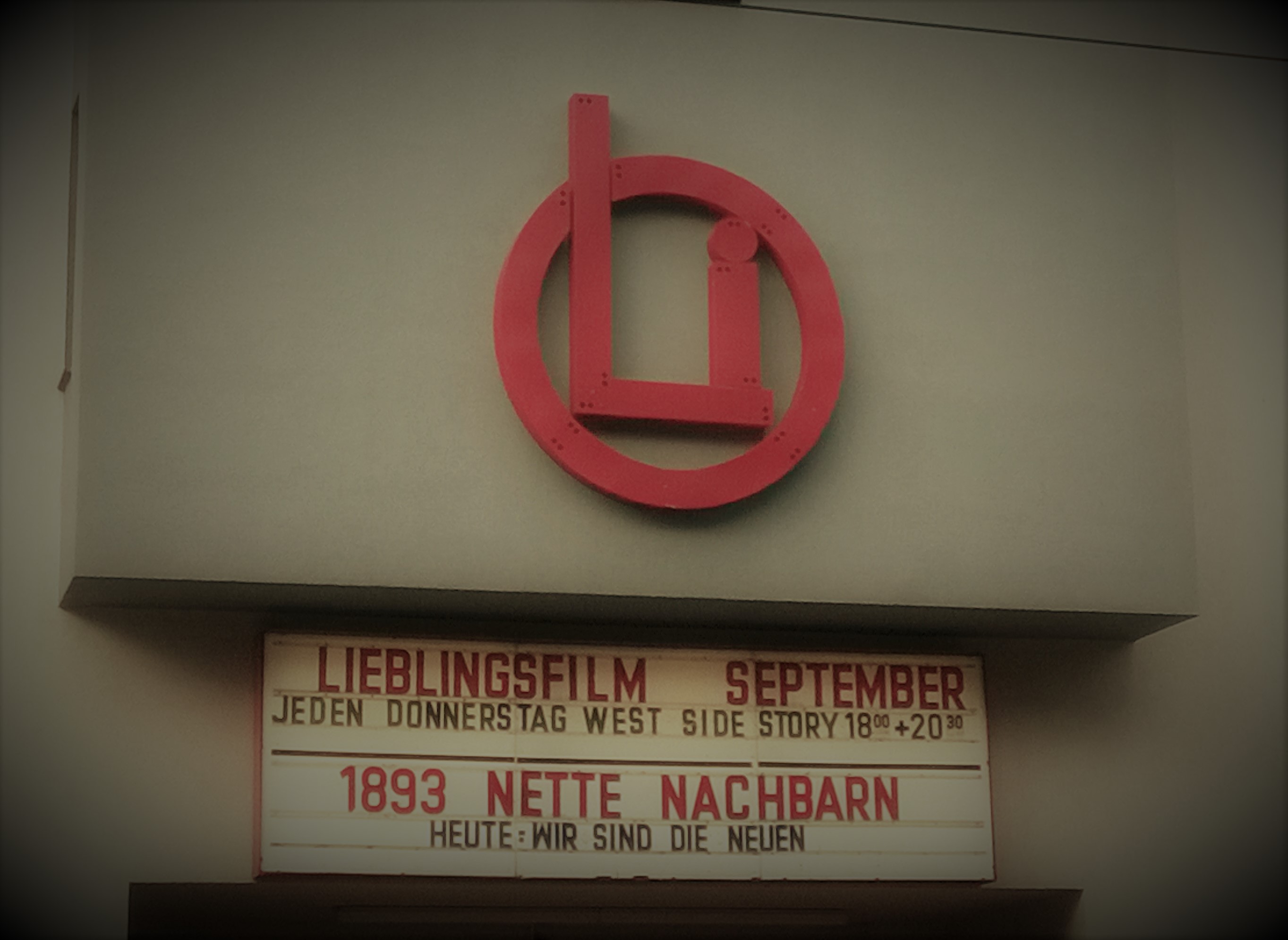 Hauswand des OLi-Kinos mit dem roten Logo. Darunter eine alte Filmtafel mit der Aufschrift "1893 Nette Nachbarn"