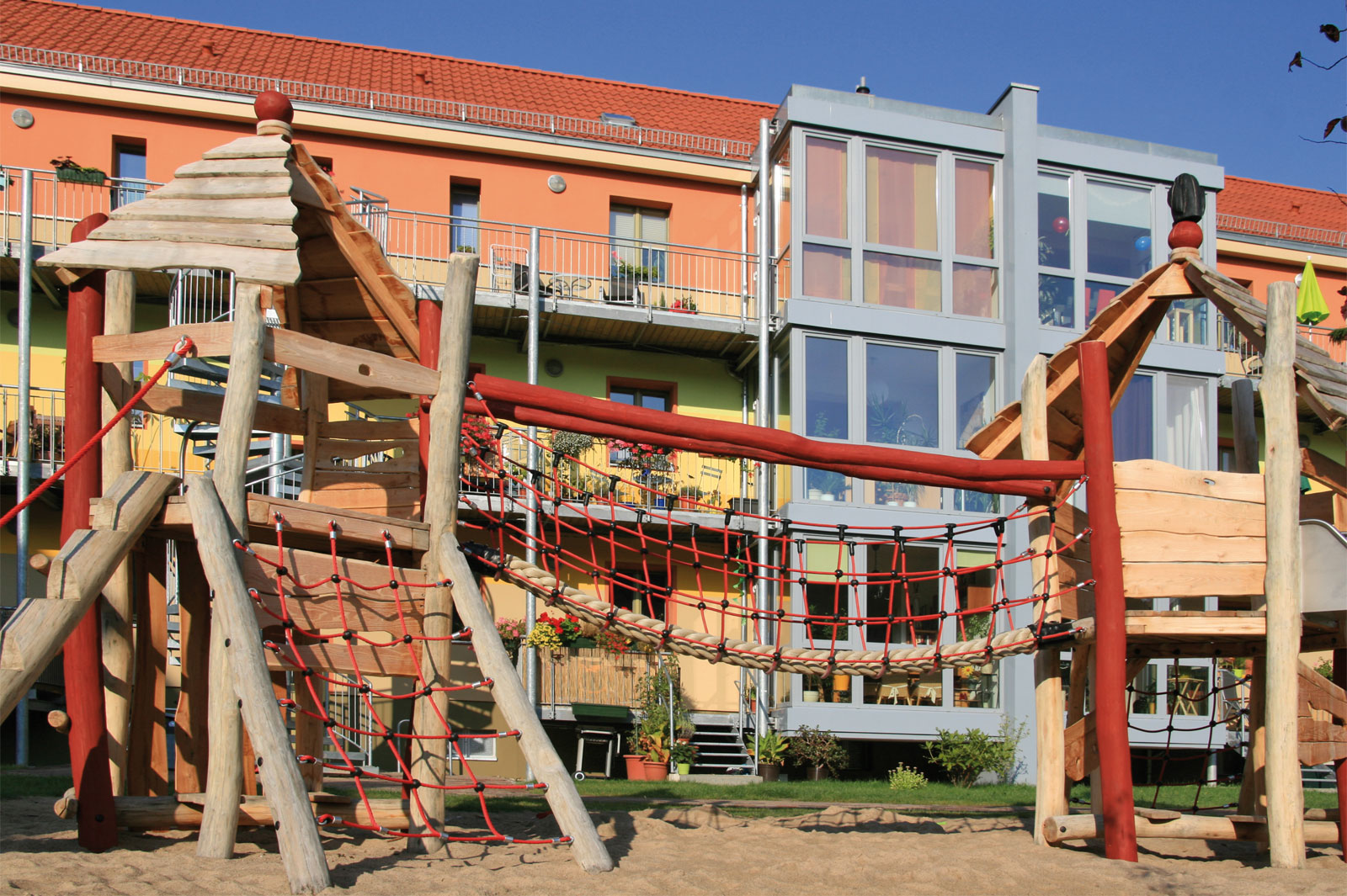 Holz-Klettergerüst mit Netzbrücke auf einem Spielplatz vor buntem Wohngebäude