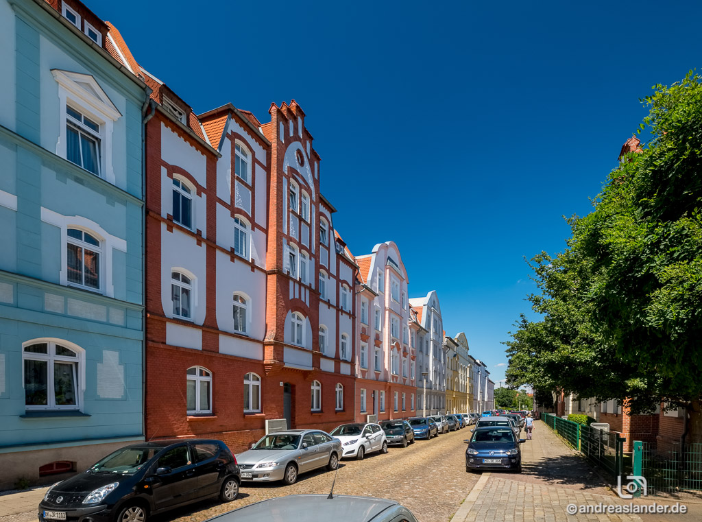 Reihe historischer Wohnhäuser mit roten und blauen Fassaden an einer ruhigen Straße unter blauem Himmel